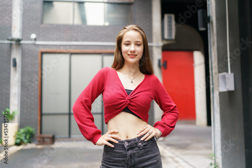 Stylish Young Woman in Urban Setting: A confident young woman with a bright smile stands in an urban alleyway, posing casually with her hands on her hips, showcasing her fashionable attire.