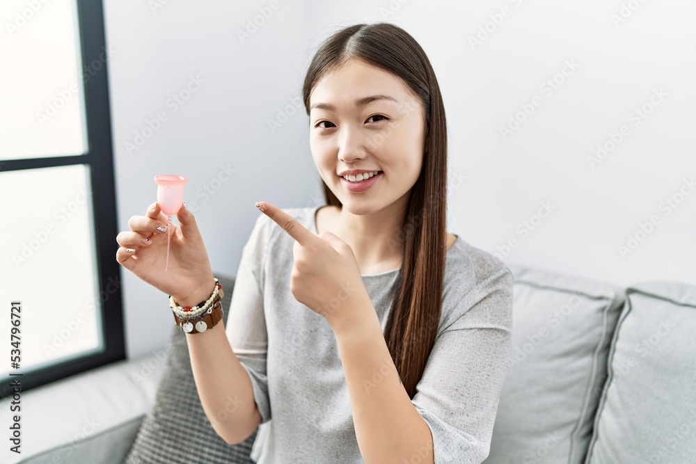 Young asian woman holding menstrual cup sitting on the sofa smiling happy pointing with hand and finger Young asian woman holding menstrual cup sitting on the sofa smiling happy pointing with hand and finger