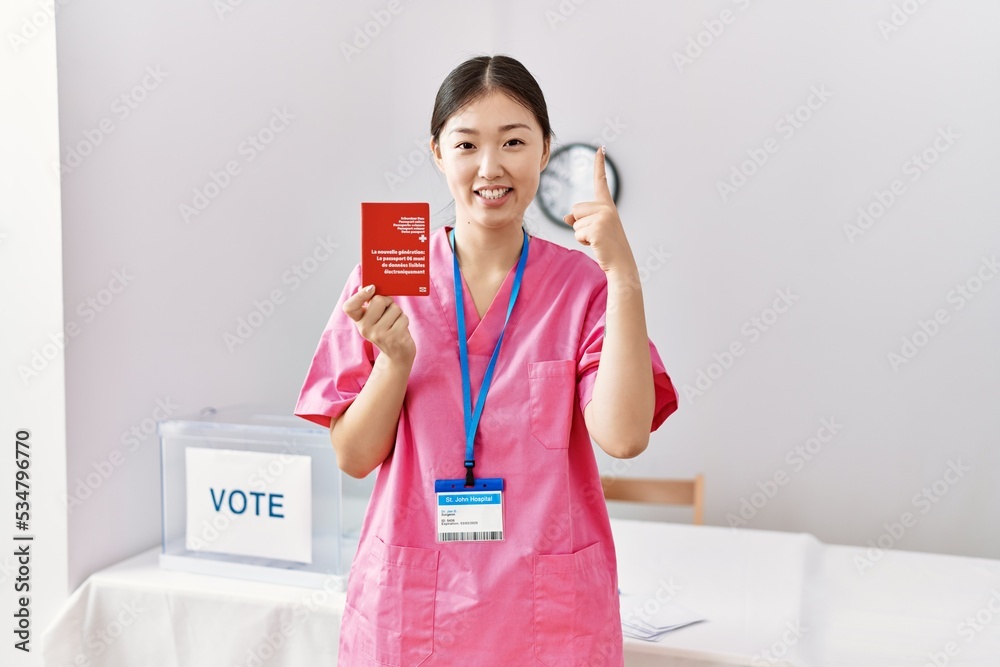 Young asian nurse woman at political campaign election holding swiss passport smiling with an idea or question pointing finger with happy face, number one Young asian nurse woman at political campaign election holding swiss passport smiling with an idea or question pointing finger with happy face, number one