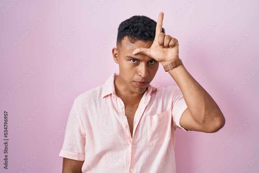 Young hispanic man standing over pink background making fun of people with fingers on forehead doing loser gesture mocking and insulting.