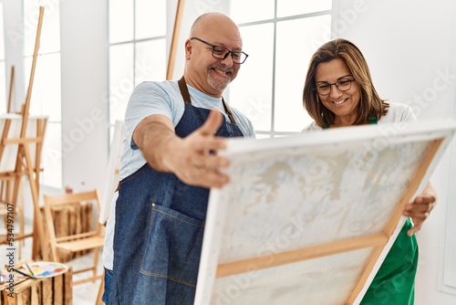 Middle age hispanic painter couple smiling happy looking canvas at art studio.