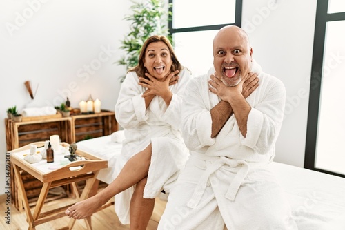 Middle age hispanic couple wearing bathrobe at wellness spa shouting and suffocate because painful strangle. health problem. asphyxiate and suicide concept.