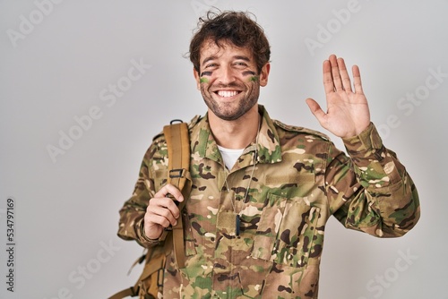 Hispanic young man wearing camouflage army uniform waiving saying hello happy and smiling, friendly welcome gesture