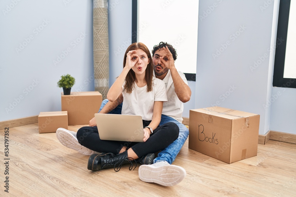 Young couple using laptop at new home doing ok gesture shocked with surprised face, eye looking through fingers. unbelieving expression. Young couple using laptop at new home doing ok gesture shocked with surprised face, eye looking through fingers. unbelieving expression.
