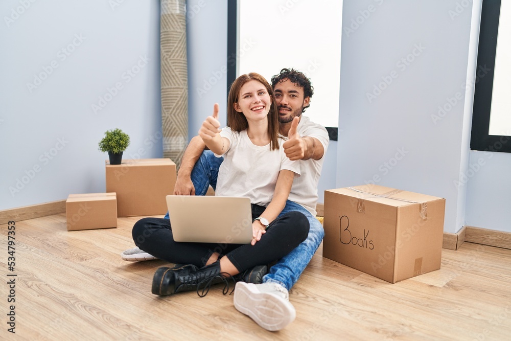 Young couple using laptop at new home approving doing positive gesture with hand, thumbs up smiling and happy for success. winner gesture. Young couple using laptop at new home approving doing positive gesture with hand, thumbs up smiling and happy for success. winner gesture.