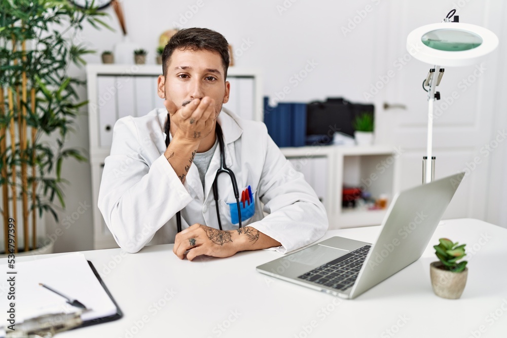 Young doctor working at the clinic using computer laptop looking at the camera blowing a kiss with hand on air being lovely and sexy. love expression.