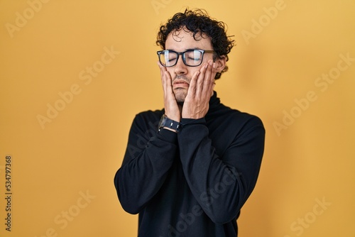 Hispanic man standing over yellow background tired hands covering face, depression and sadness, upset and irritated for problem