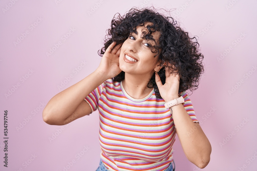 Young middle east woman standing over pink background trying to hear both hands on ear gesture, curious for gossip. hearing problem, deaf
