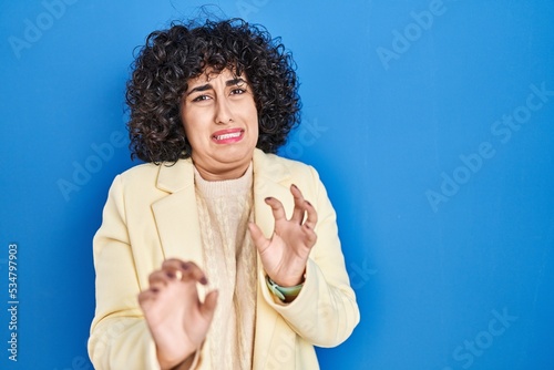 Young brunette woman with curly hair standing over blue background disgusted expression, displeased and fearful doing disgust face because aversion reaction.