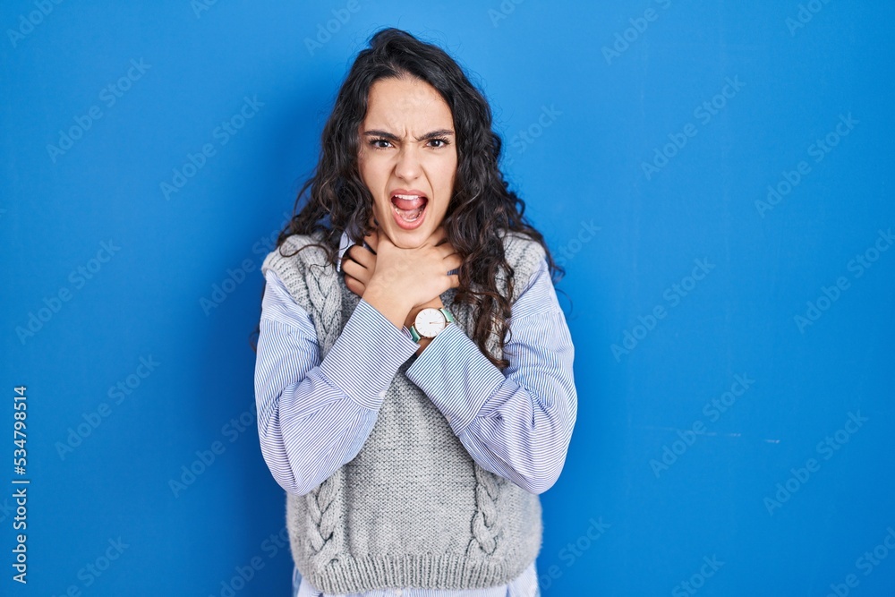 Obraz premium Young brunette woman standing over blue background shouting suffocate because painful strangle. health problem. asphyxiate and suicide concept.