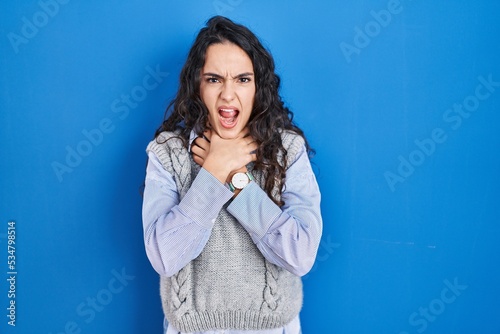 Young brunette woman standing over blue background shouting suffocate because painful strangle. health problem. asphyxiate and suicide concept.