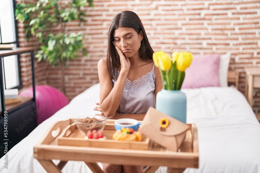 Brunette young woman eating breakfast sitting on the bed thinking looking tired and bored with depression problems with crossed arms.