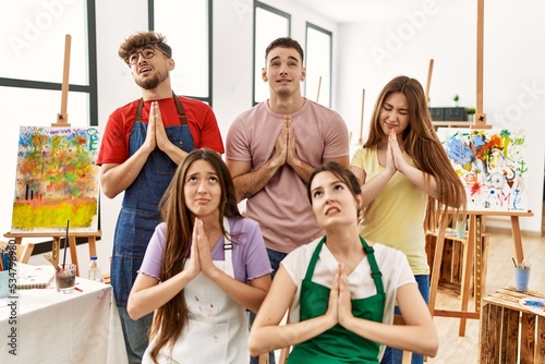 Group of five hispanic artists at art studio begging and praying with hands together with hope expression on face very emotional and worried. begging.