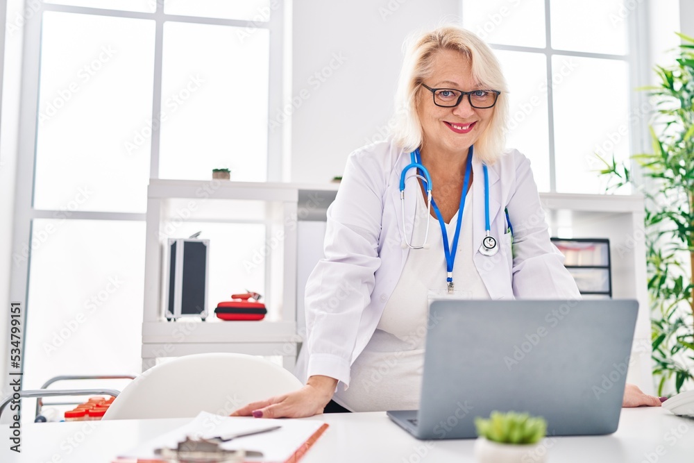 Middle age blonde woman wearing doctor uniform using laptop at clinic Middle age blonde woman wearing doctor uniform using laptop at clinic