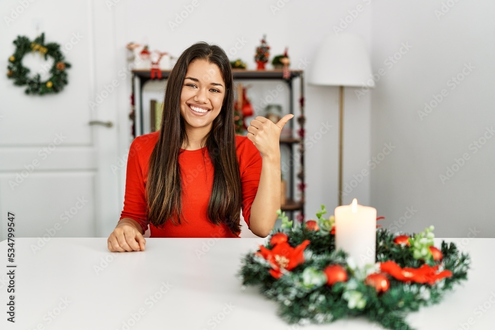 Young brunette woman sitting on the table on christmas smiling with happy face looking and pointing to the side with thumb up. Young brunette woman sitting on the table on christmas smiling with happy face looking and pointing to the side with thumb up.