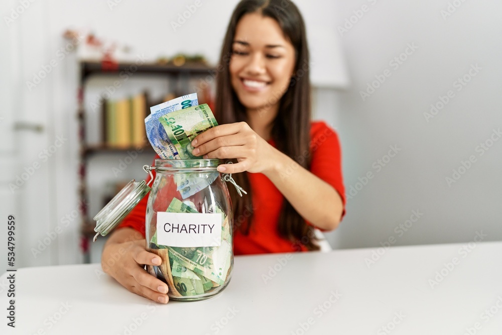 Young latin woman holding charity jar with rands banknotes sitting by christmas decor at home Young latin woman holding charity jar with rands banknotes sitting by christmas decor at home