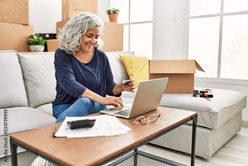 Middle age grey-haired woman sitting on the sofa using laptop and credit card to buy at new home.