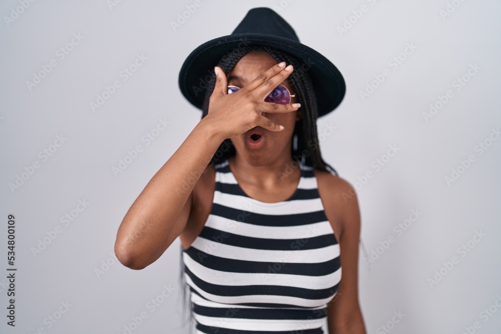 Young african american with braids wearing hat and sunglasses peeking in shock covering face and eyes with hand, looking through fingers with embarrassed expression. Young african american with braids wearing hat and sunglasses peeking in shock covering face and eyes with hand, looking through fingers with embarrassed expression.