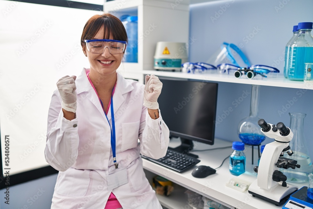 Young brunette woman working at scientist laboratory excited for success with arms raised and eyes closed celebrating victory smiling. winner concept. Young brunette woman working at scientist laboratory excited for success with arms raised and eyes closed celebrating victory smiling. winner concept.