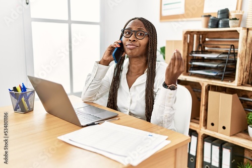 Black woman with braids working at the office speaking on the phone doing italian gesture with hand and fingers confident expression