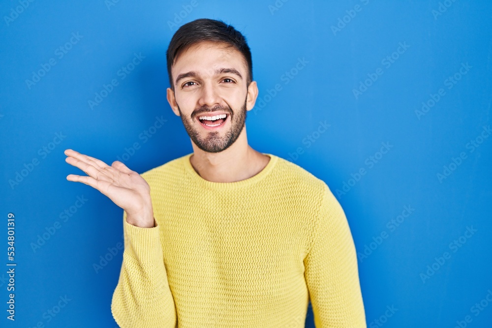 Hispanic man standing over blue background smiling cheerful presenting and pointing with palm of hand looking at the camera. Hispanic man standing over blue background smiling cheerful presenting and pointing with palm of hand looking at the camera.