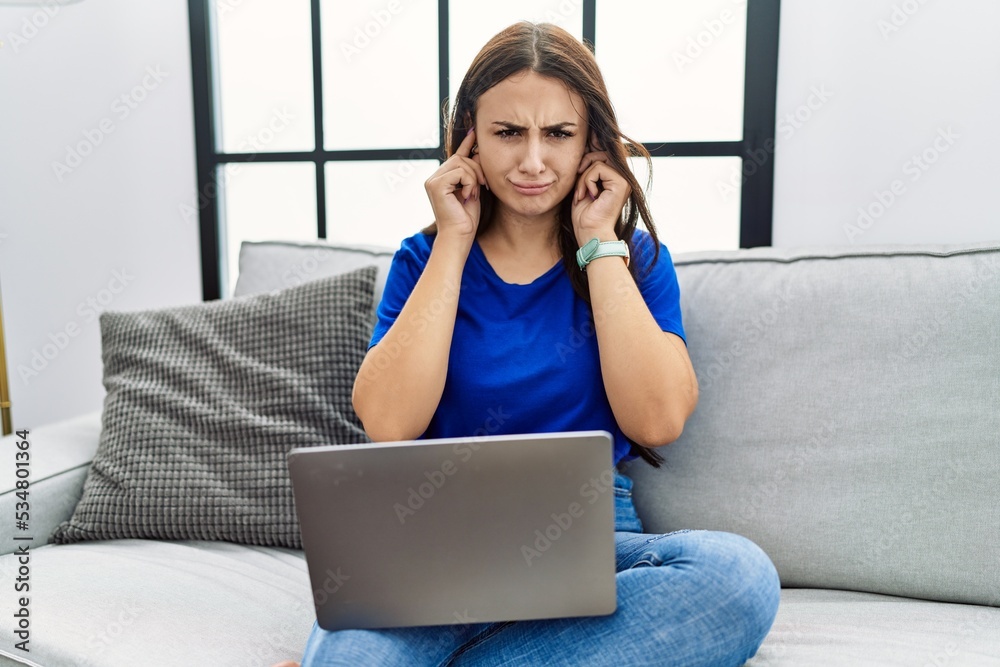 Young brunette woman using laptop at home covering ears with fingers with annoyed expression for the noise of loud music. deaf concept.