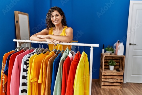 Young latin woman smiling confident leaning on clothes rack at laundry room