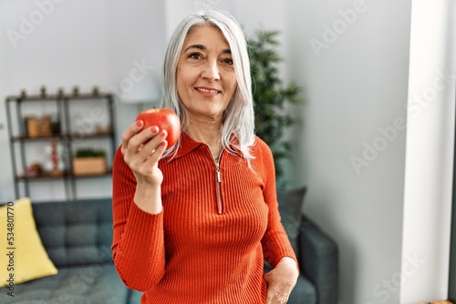 Middle age grey-haired woman smiling confident holding red apple at home