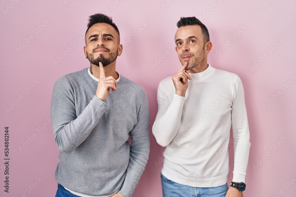 Homosexual couple standing over pink background thinking concentrated about doubt with finger on chin and looking up wondering Homosexual couple standing over pink background thinking concentrated about doubt with finger on chin and looking up wondering