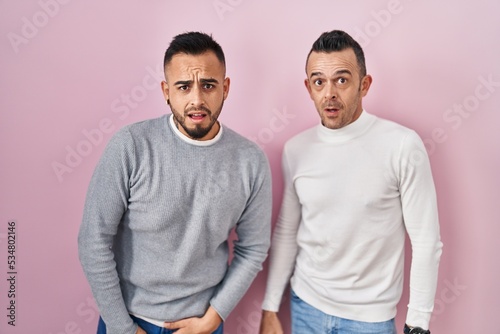 Homosexual couple standing over pink background in shock face, looking skeptical and sarcastic, surprised with open mouth