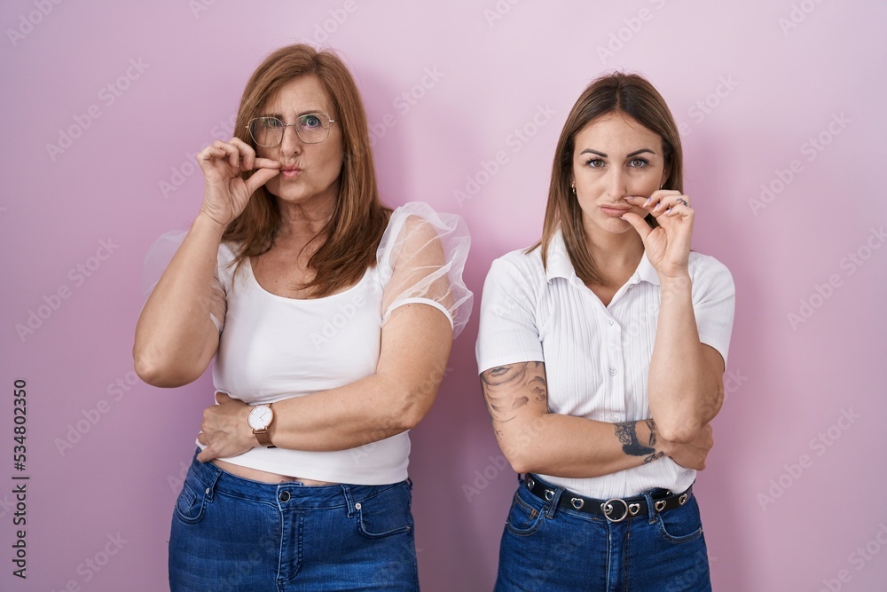 Hispanic mother and daughter wearing casual white t shirt over pink background mouth and lips shut as zip with fingers. secret and silent, taboo talking Hispanic mother and daughter wearing casual white t shirt over pink background mouth and lips shut as zip with fingers. secret and silent, taboo talking