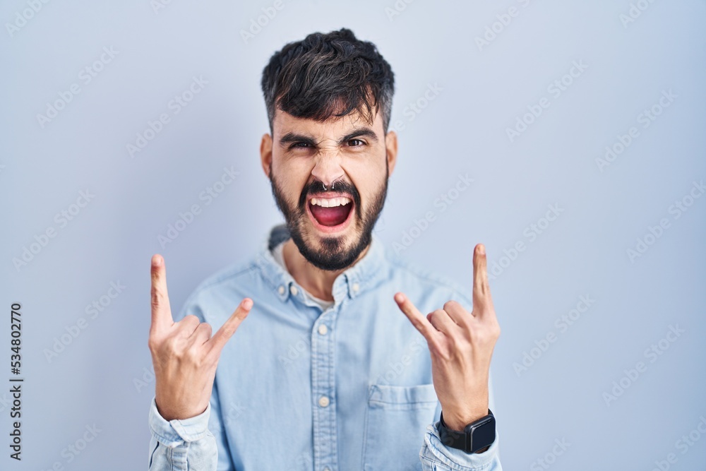 Young hispanic man with beard standing over blue background shouting with crazy expression doing rock symbol with hands up. music star. heavy music concept. Young hispanic man with beard standing over blue background shouting with crazy expression doing rock symbol with hands up. music star. heavy music concept.