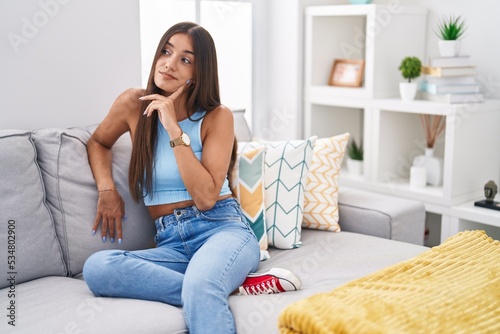 Young brunette woman sitting on the sofa at home with hand on chin thinking about question, pensive expression. smiling with thoughtful face. doubt concept.