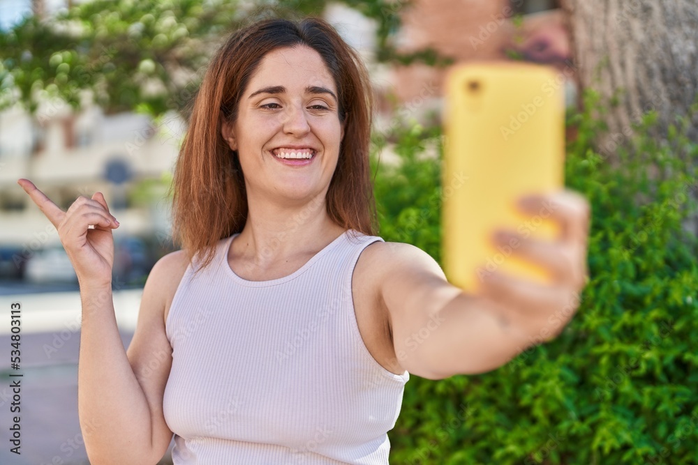 Brunette woman taking a selfie photo with smartphone smiling happy pointing with hand and finger to the side Brunette woman taking a selfie photo with smartphone smiling happy pointing with hand and finger to the side