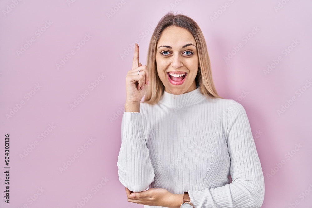Young blonde woman wearing white sweater over pink background pointing finger up with successful idea. exited and happy. number one. Young blonde woman wearing white sweater over pink background pointing finger up with successful idea. exited and happy. number one.