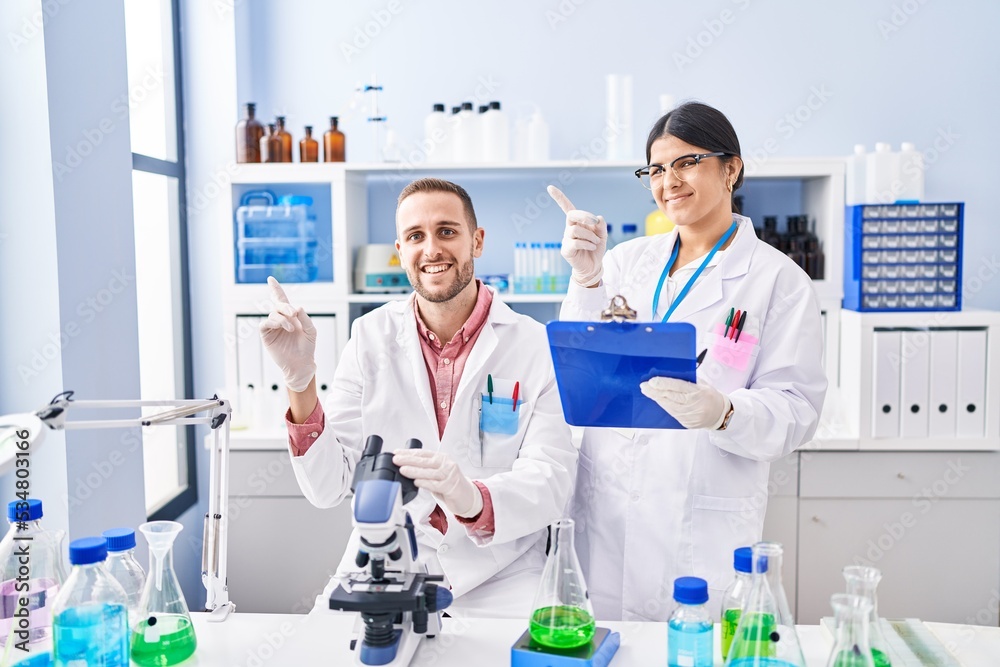 Two young people working at scientist laboratory smiling happy pointing with hand and finger to the side Two young people working at scientist laboratory smiling happy pointing with hand and finger to the side