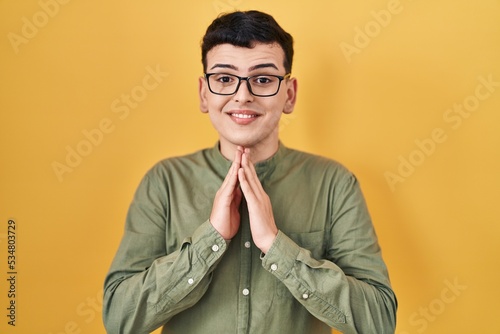 Non binary person standing over yellow background praying with hands together asking for forgiveness smiling confident.