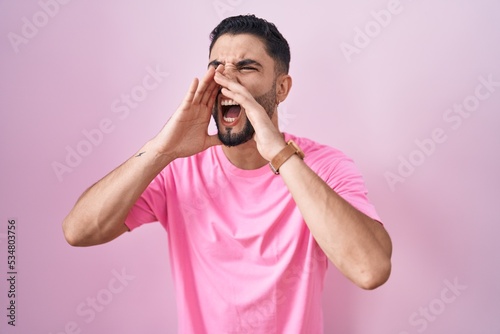 Hispanic young man standing over pink background shouting angry out loud with hands over mouth