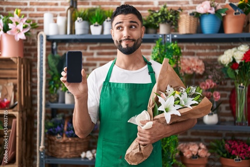 Hispanic young man working at florist shop showing smartphone screen smiling looking to the side and staring away thinking.