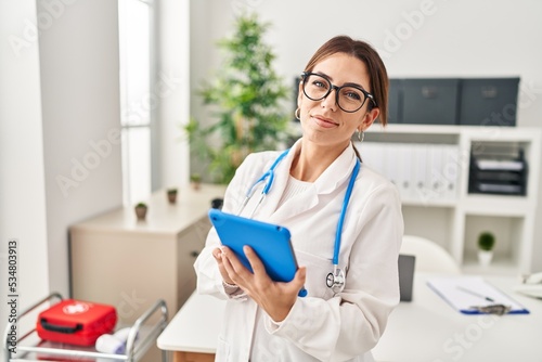 Young hispanic woman wearing doctor uniform using touchpad at clinic