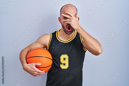 Young bald man with beard wearing basketball uniform holding ball peeking in shock covering face and eyes with hand, looking through fingers with embarrassed expression.