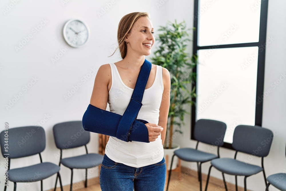 Young caucasian woman wearing sling on arm for accident standing at clinic waiting room