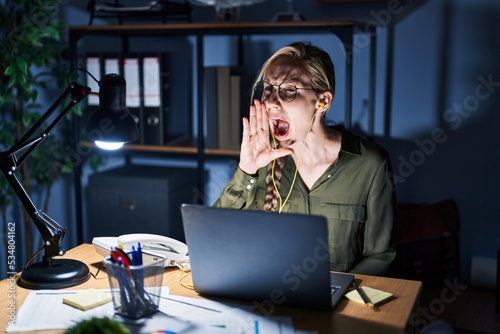 Young blonde woman working at the office at night shouting and screaming loud to side with hand on mouth. communication concept.