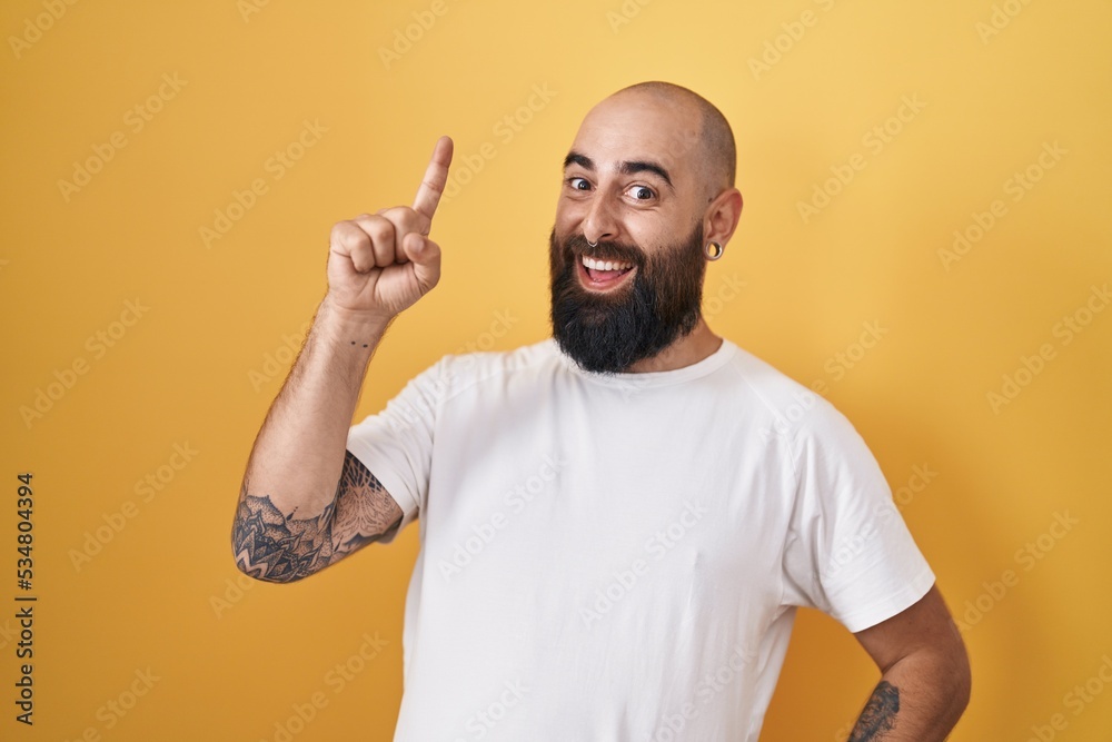 Young hispanic man with beard and tattoos standing over yellow background pointing finger up with successful idea. exited and happy. number one. Young hispanic man with beard and tattoos standing over yellow background pointing finger up with successful idea. exited and happy. number one.