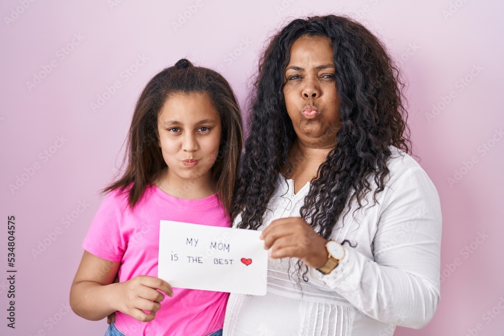 Mother and young daughter holding my mom is the best banner puffing cheeks with funny face. mouth inflated with air, catching air. Mother and young daughter holding my mom is the best banner puffing cheeks with funny face. mouth inflated with air, catching air.
