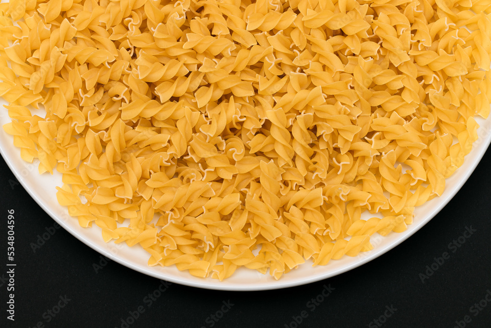 Uncooked Fusilli Pasta Lying on White Plate on Black Background. Raw and Dry Macaroni. Unhealthy and Fat Food - Top View Uncooked Fusilli Pasta Lying on White Plate on Black Background. Raw and Dry Macaroni. Unhealthy and Fat Food - Top View