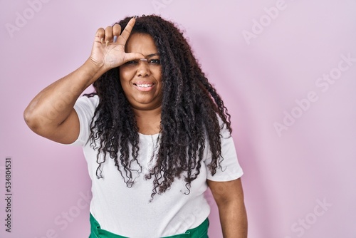 Plus size hispanic woman standing over pink background making fun of people with fingers on forehead doing loser gesture mocking and insulting. photo