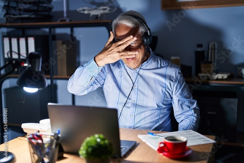 Hispanic senior man wearing call center agent headset at night peeking in shock covering face and eyes with hand, looking through fingers with embarrassed expression.