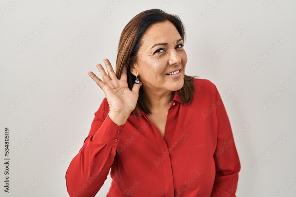 Hispanic mature woman standing over white background smiling with hand over ear listening an hearing to rumor or gossip. deafness concept. Hispanic mature woman standing over white background smiling with hand over ear listening an hearing to rumor or gossip. deafness concept.