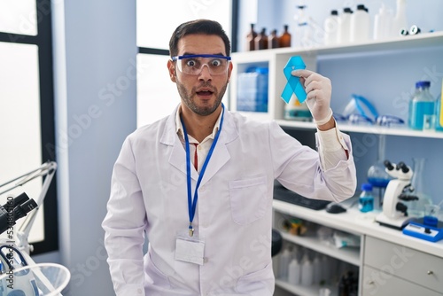 Young hispanic man with beard working at scientist laboratory holding blue ribbon scared and amazed with open mouth for surprise, disbelief face